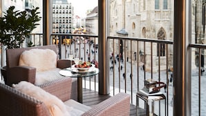 A balcony with wicker furniture, a table with a fruit bowl and glasses, and a view of a historic building and busy street. - DO & CO Hotel Vienna (Vienna)