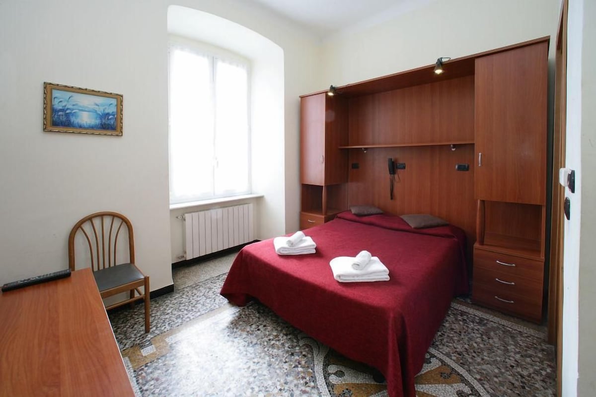 A bedroom with a bed, a wooden wardrobe, a chair, a desk, and a painting on the wall.
