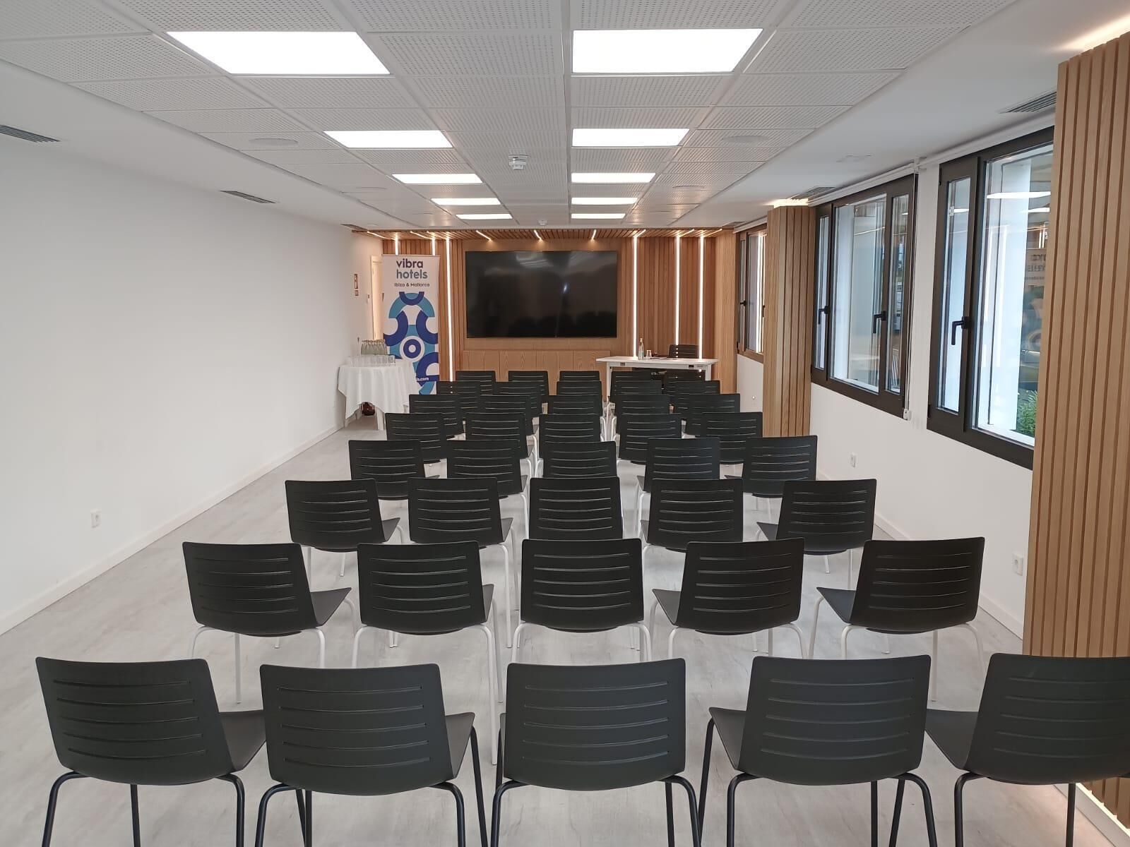 A conference room with rows of black chairs, a large screen, and a wooden panel wall.