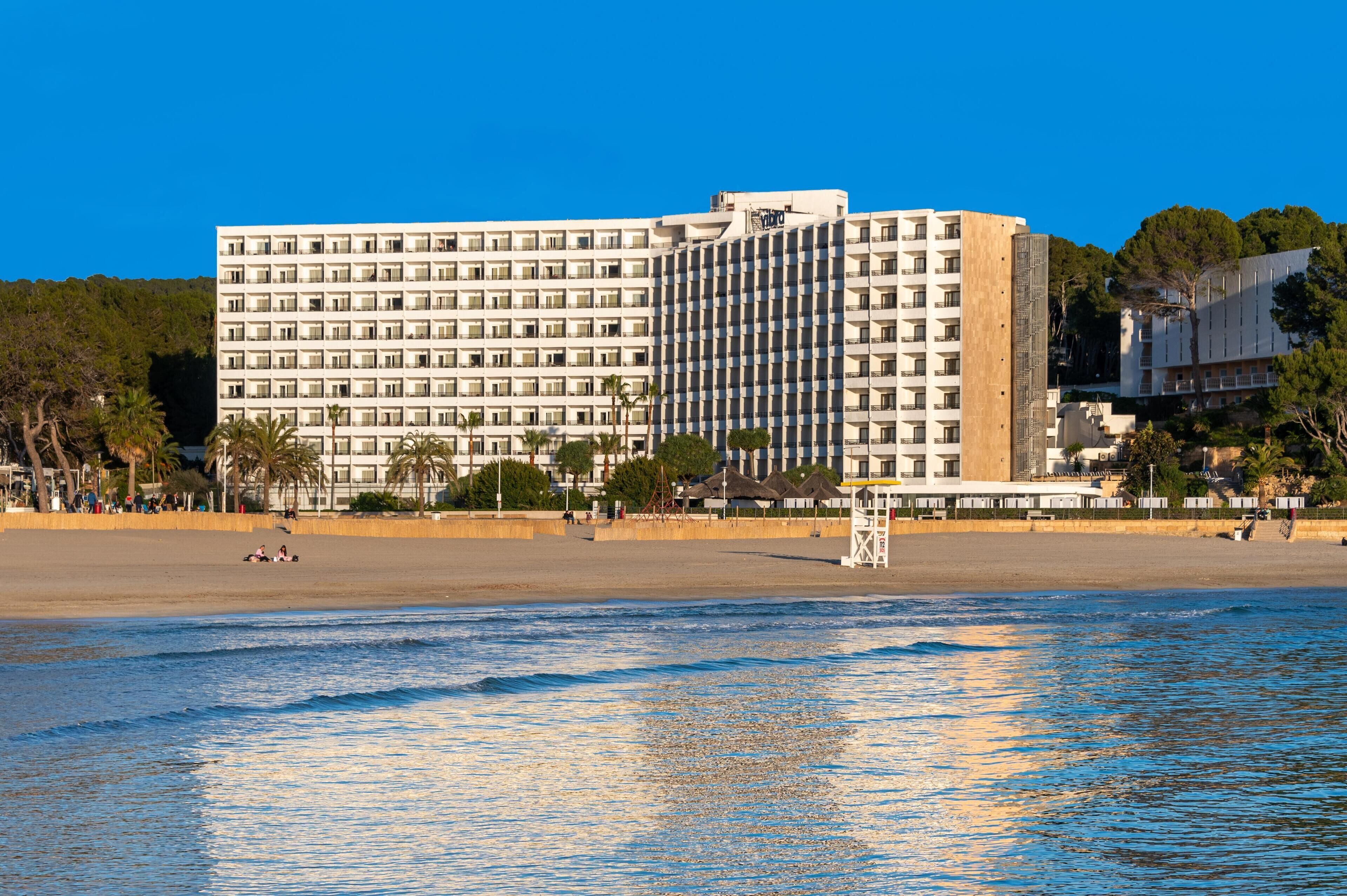 Een groot hotel met meerdere verdiepingen en balkons, gelegen aan een strand met palmbomen en een badwachterspost.