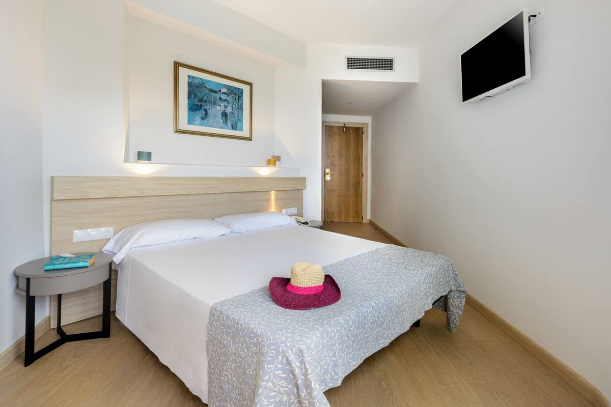 A hotel room with a bed, a bedside table with a book, a wall-mounted TV, and a door leading to another room.
