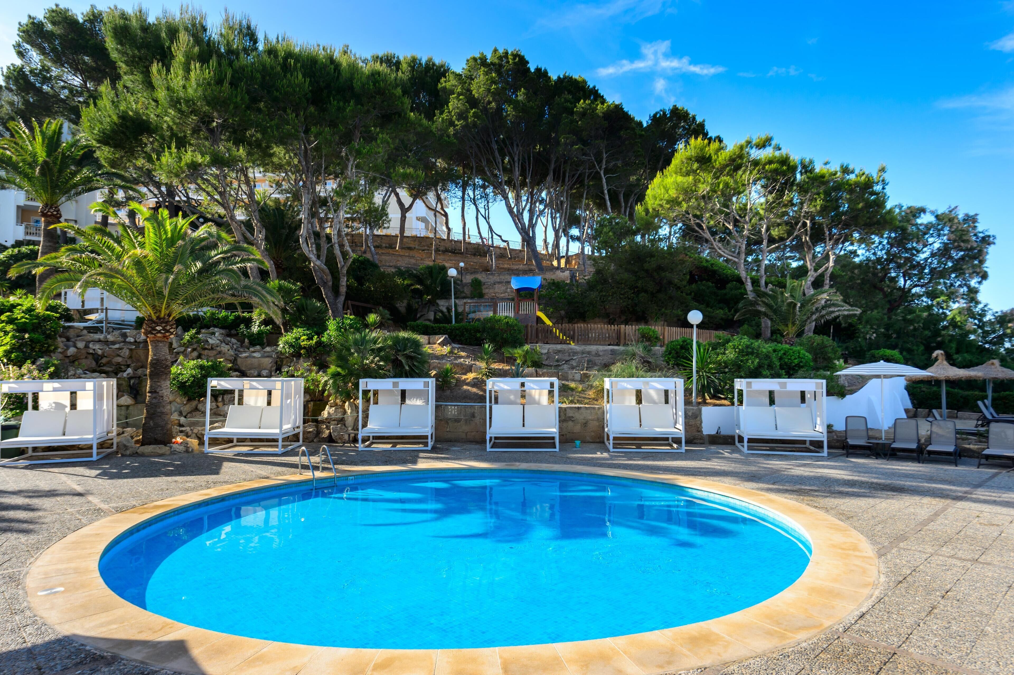 Una piscina con sillas de descanso y sombrillas, rodeada de árboles y un muro de piedra.