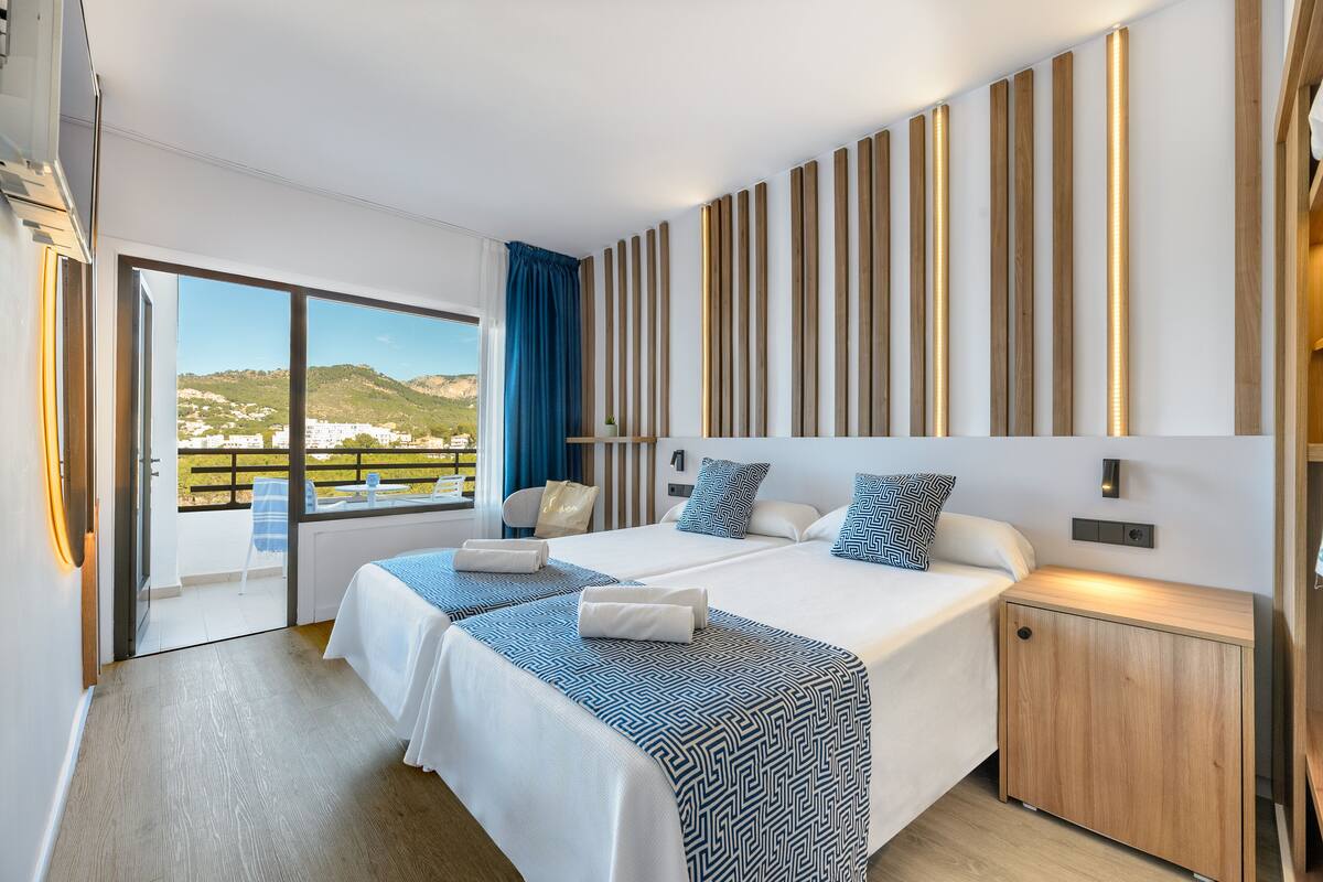 A modern hotel room with two beds, a wooden headboard, a small wooden nightstand, and a view of a balcony with a scenic landscape.