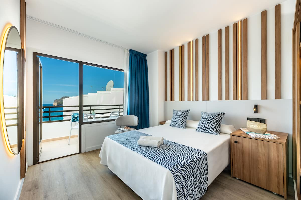 A modern hotel room with a large bed, a wooden nightstand, a mirror, and a view of the ocean.