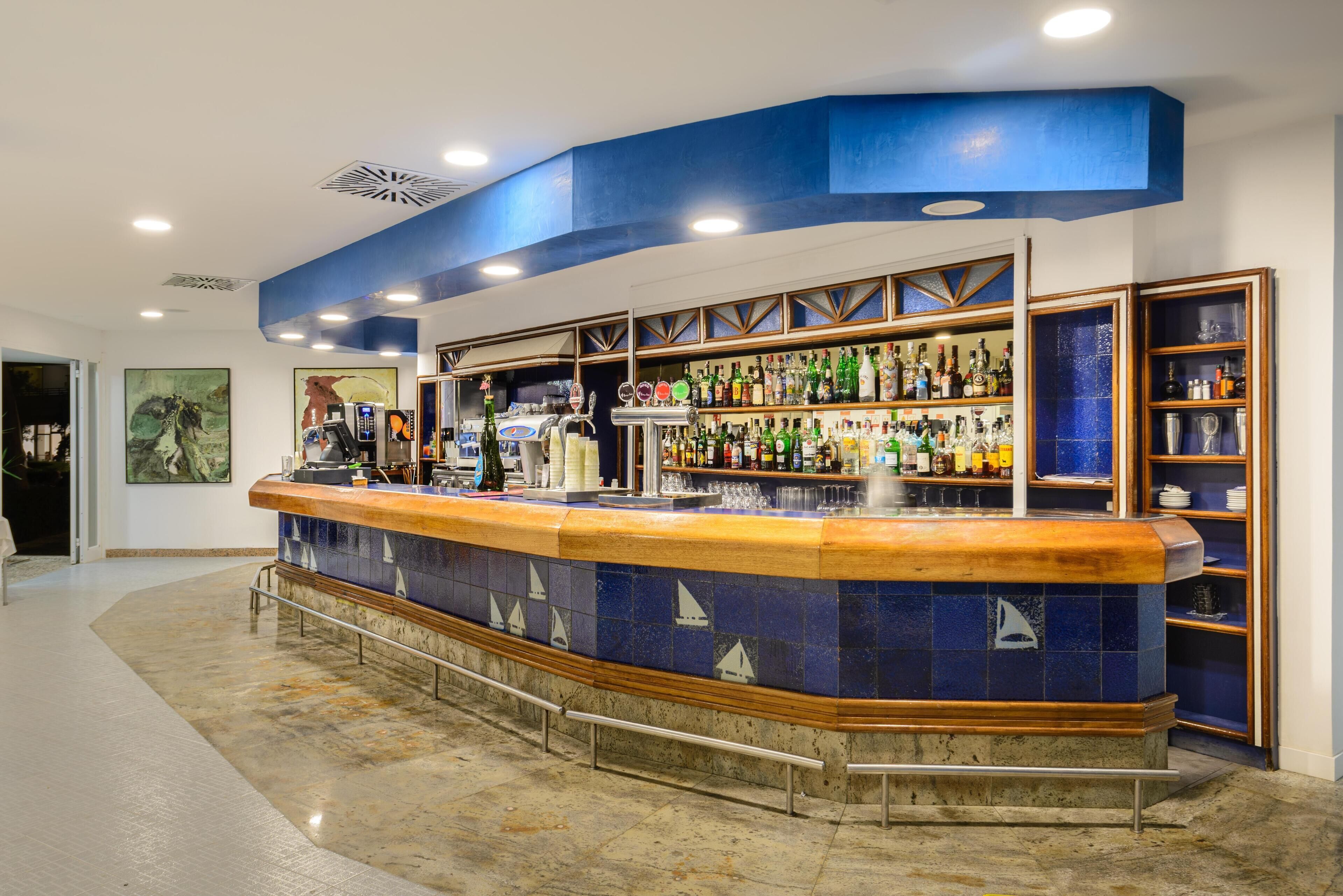 Zona de bar con mostrador curvo, estantes con botellas y pared de azulejos con diseños de veleros.