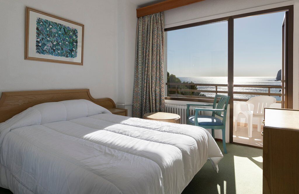 A hotel room with a large bed, a small table, a chair, and a view of the sea through a balcony.