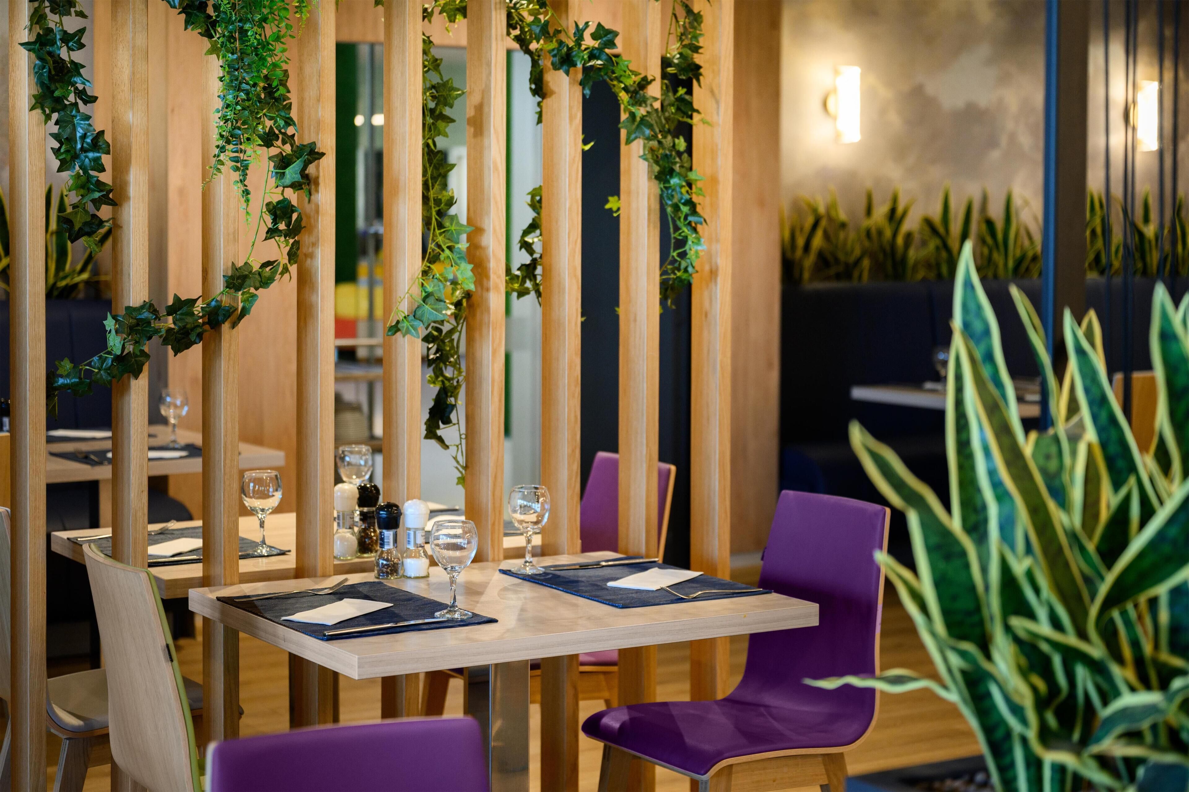 Un restaurante con divisiones verticales de madera, follaje verde, sillas moradas y una mesa con vasos y cubiertos.