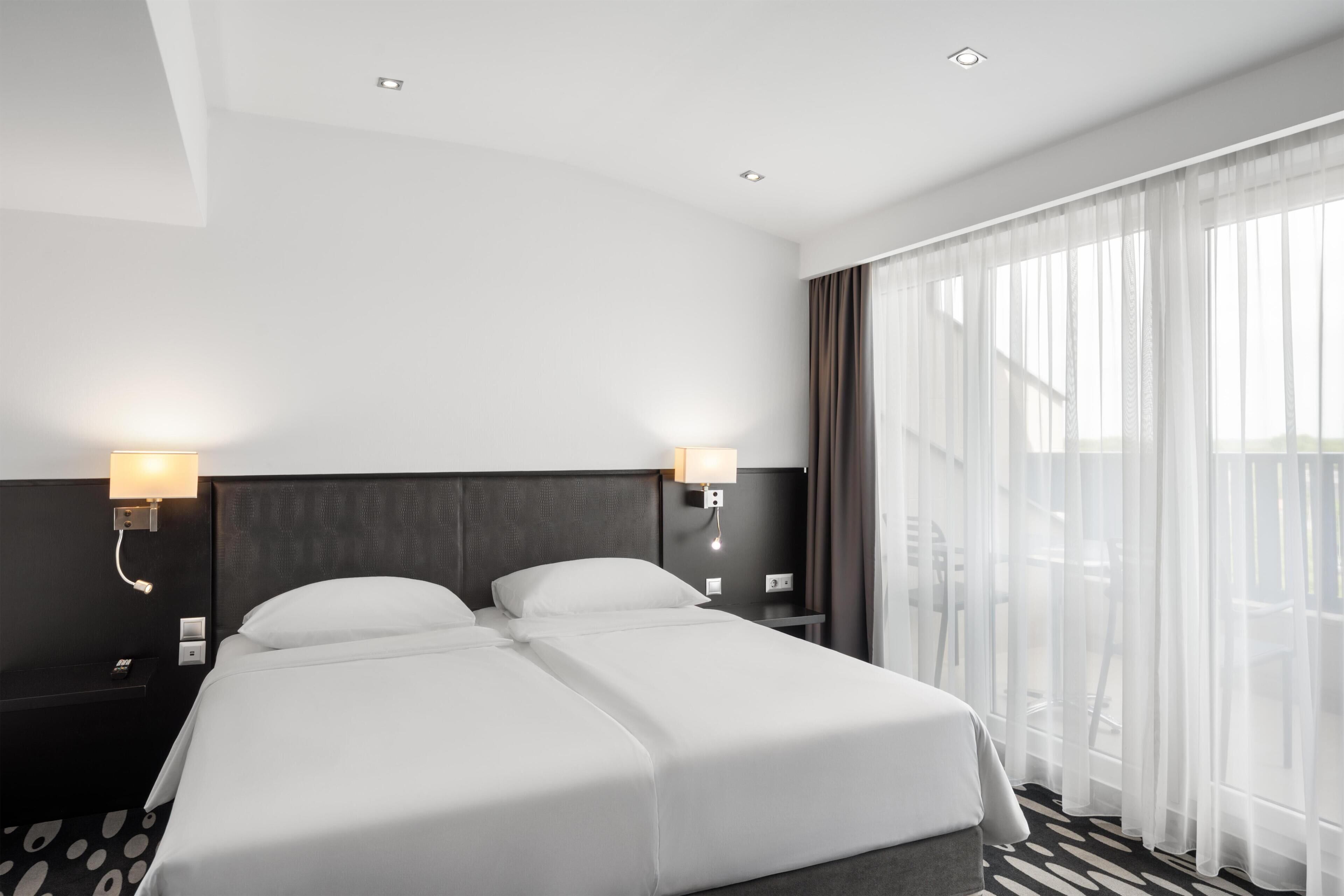 Ein Hotelzimmer mit zwei Betten, einem großen Fenster mit durchsichtigen Vorhängen, einem Nachttisch mit Lampe und einem gemusterten Teppich.