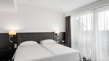 Ein Hotelzimmer mit zwei Betten, einem großen Fenster mit durchsichtigen Vorhängen, einem Nachttisch mit Lampe und einem gemusterten Teppich.