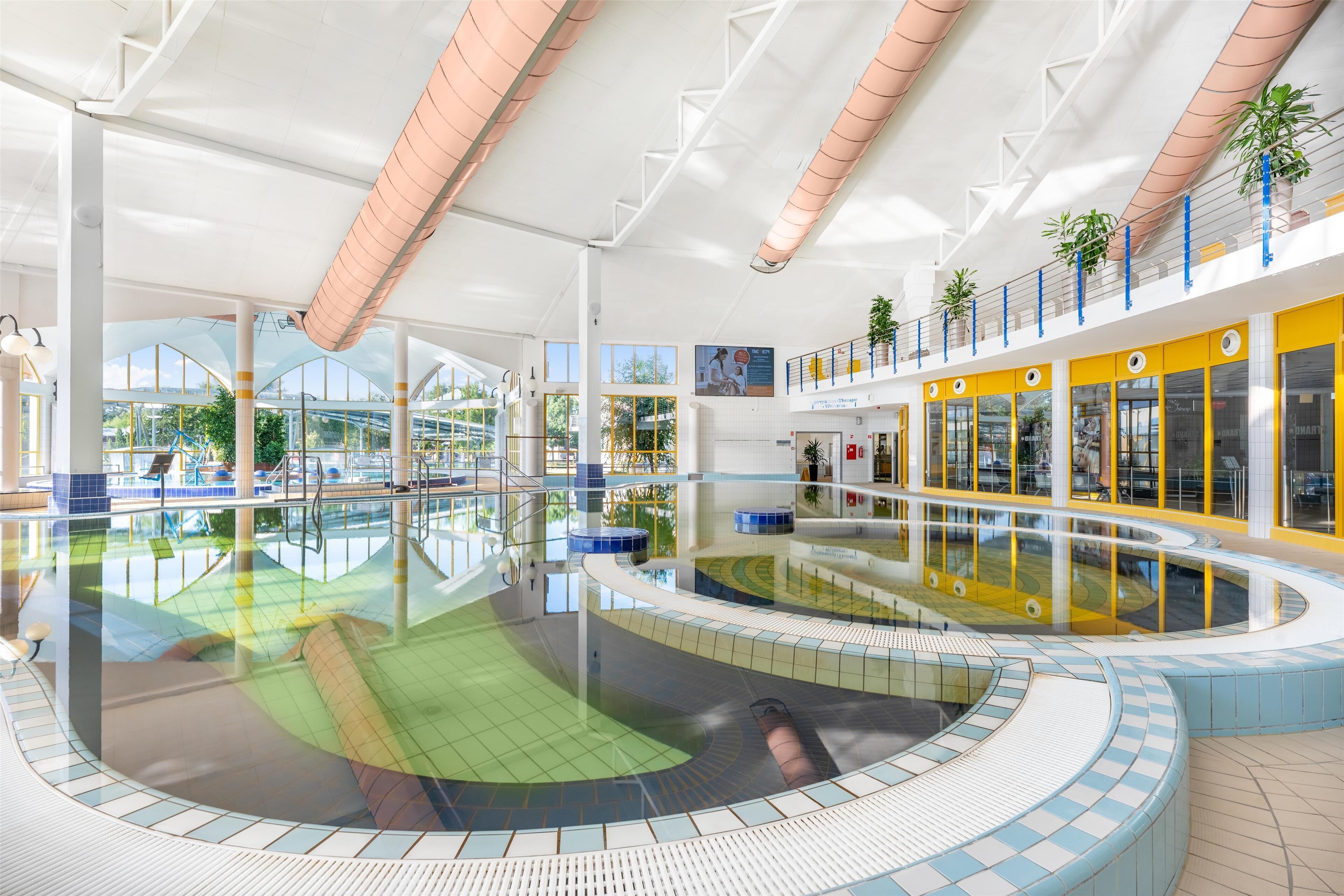 Una piscina interior moderna con una zona circular central, rodeada por un solárium con baldosas y una pared de vidrio que ofrece vistas al exterior.