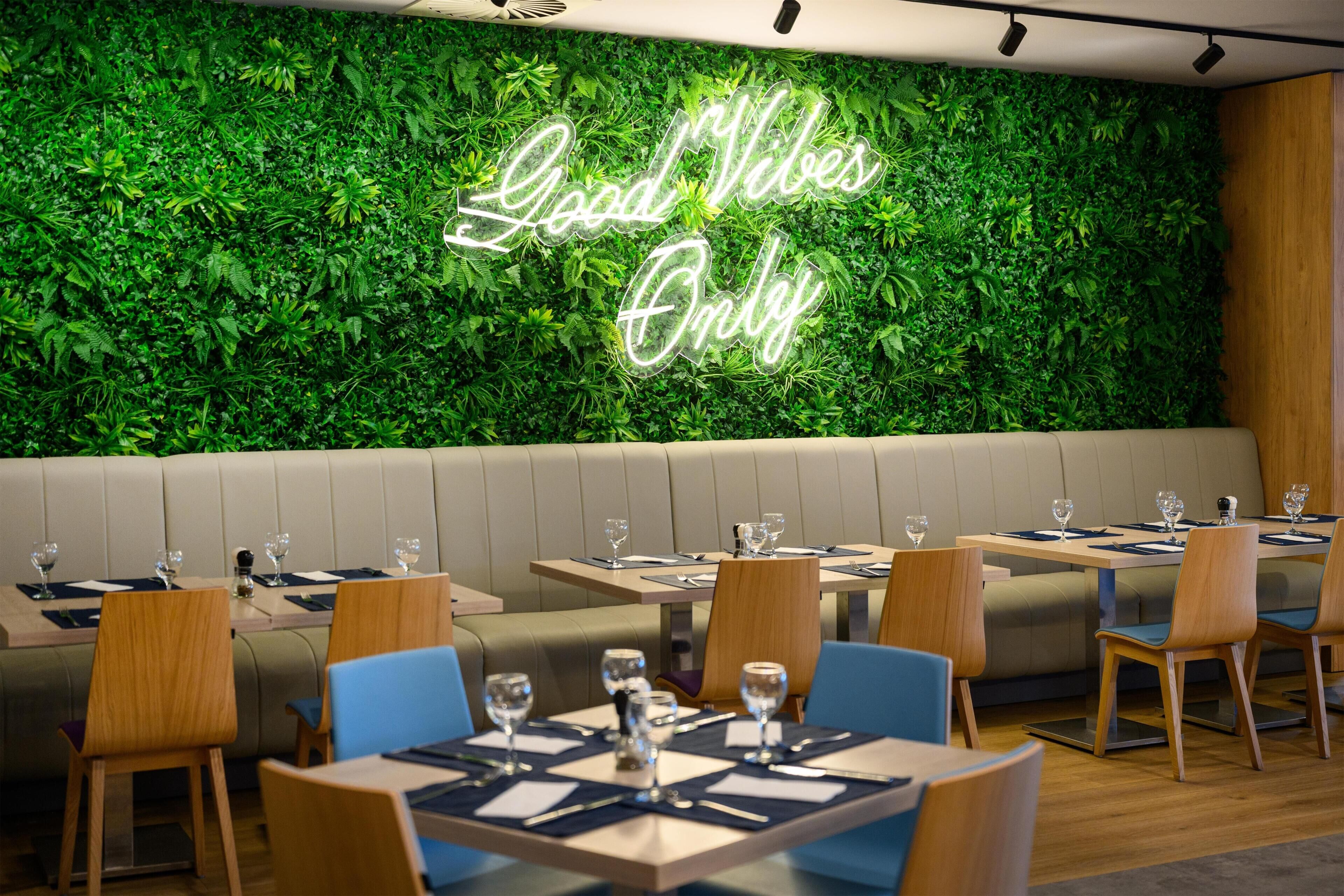 A restaurant with a neon sign reading 'Good Vibes Only' against a green leafy wall, wooden tables, and blue chairs.