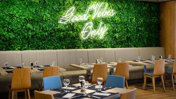 A restaurant with a neon sign reading 'Good Vibes Only' against a green leafy wall, wooden tables, and blue chairs.