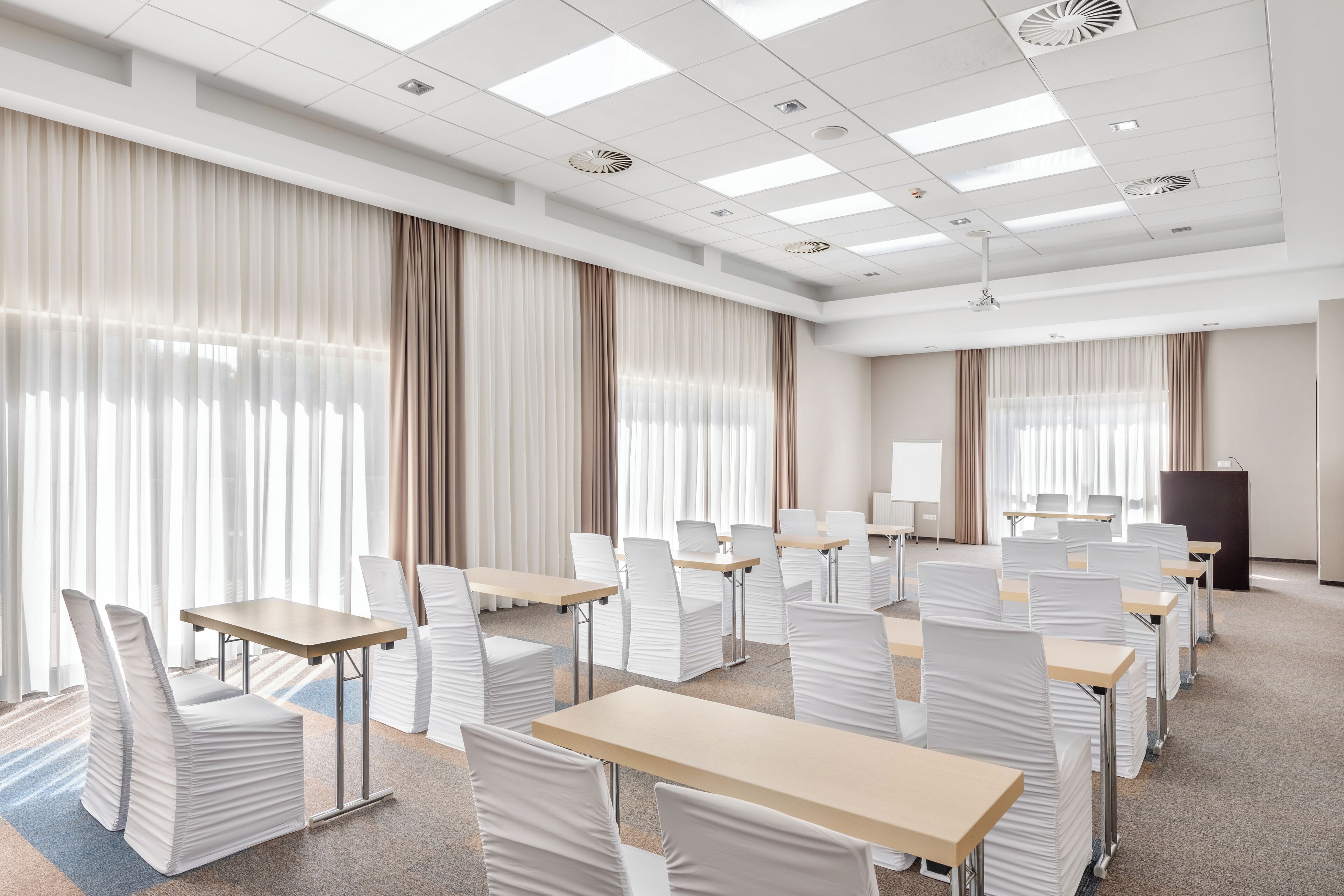 A conference room with rows of tables and chairs, large windows with curtains, and a podium.