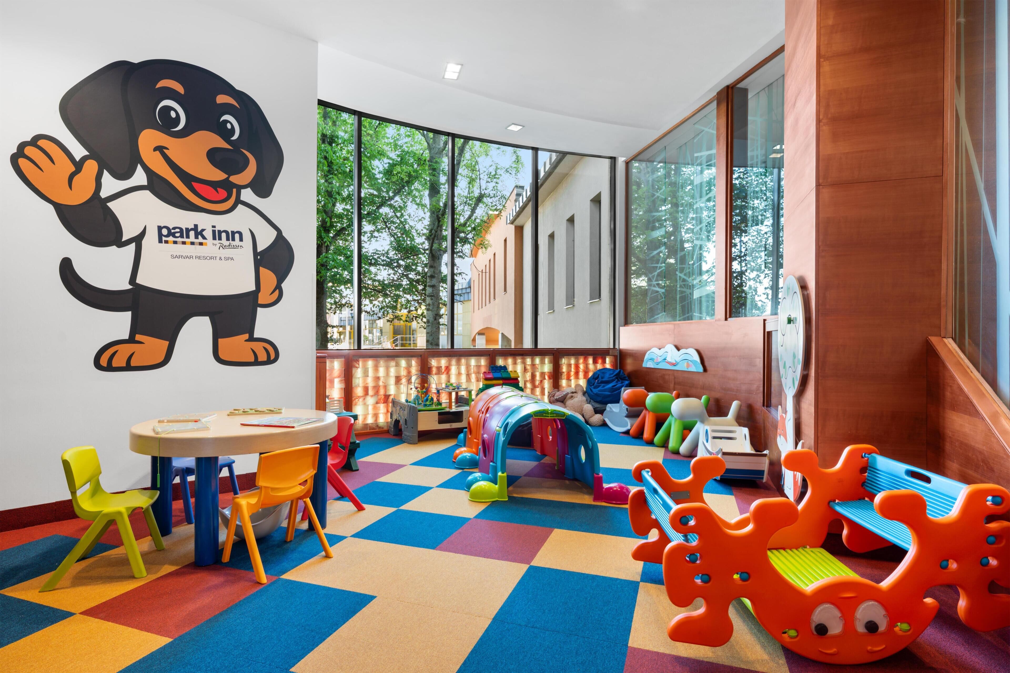 Un área de juegos infantiles con piso colorido, un gran adhesivo mural de un perro caricaturesco y vistas al exterior a través de amplias ventanas.