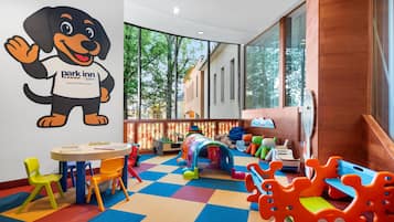 Un área de juegos infantiles con piso colorido, un gran adhesivo mural de un perro caricaturesco y vistas al exterior a través de amplias ventanas.