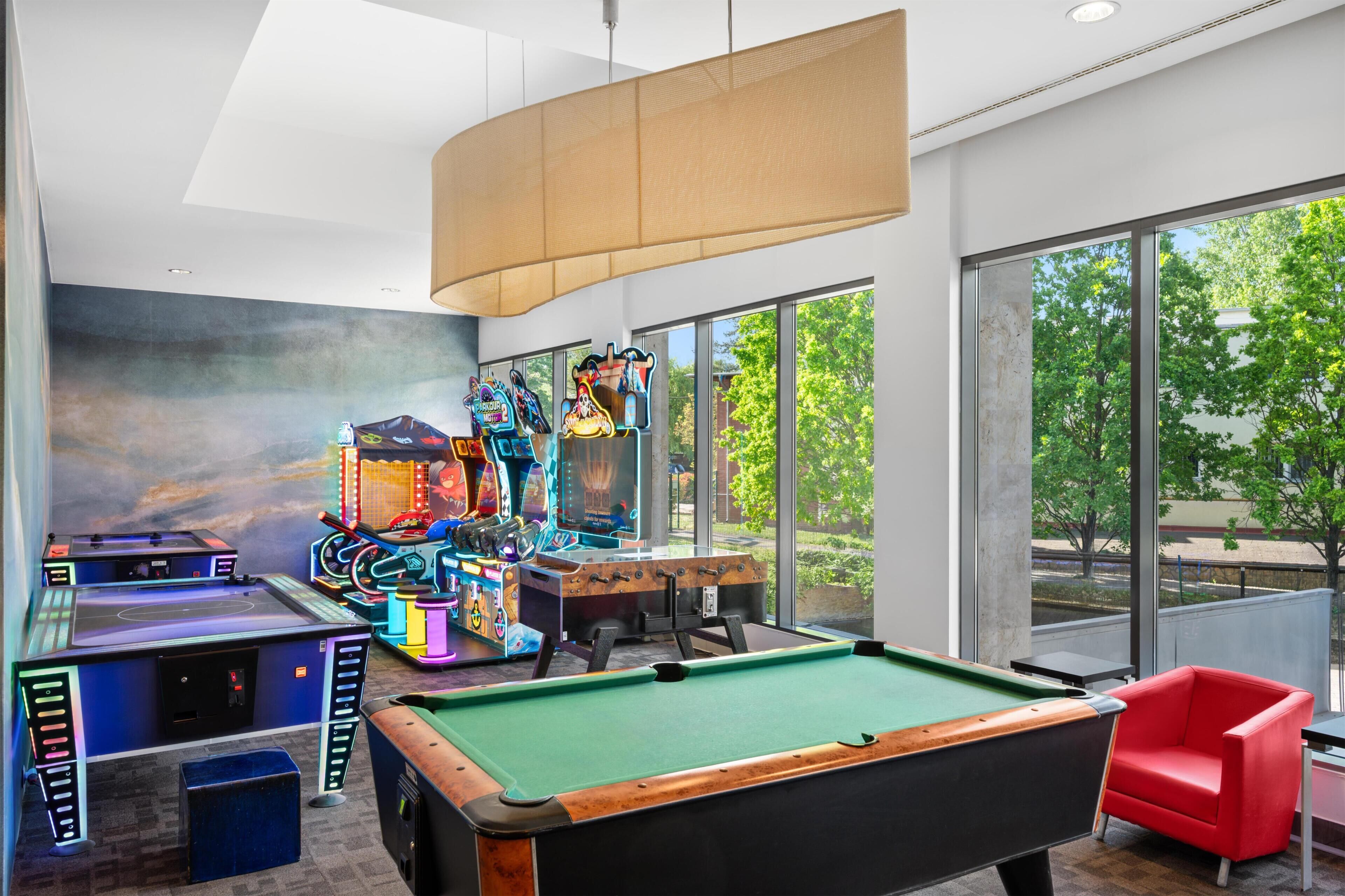 A pool table, arcade games, and a red chair in a room with large windows.