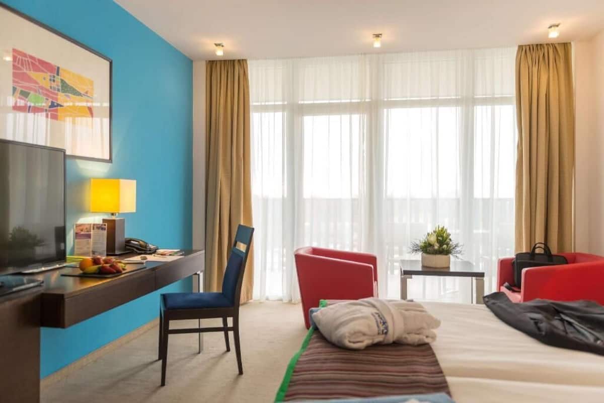 Hotellihuone, jossa on sänky, työpöytä tietokoneella, televisio ja kaksi punaista tuolia.