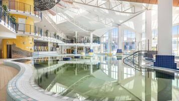A large indoor swimming pool with multiple levels and a glass ceiling, surrounded by various amenities and shops.