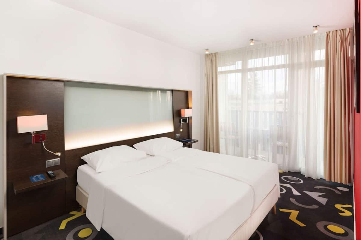 Hotellihuone, jossa on suuri sänky, yöpöytä lampun kanssa, ohjauspaneeli ja näkymä ulos ohuiden verhojen läpi.