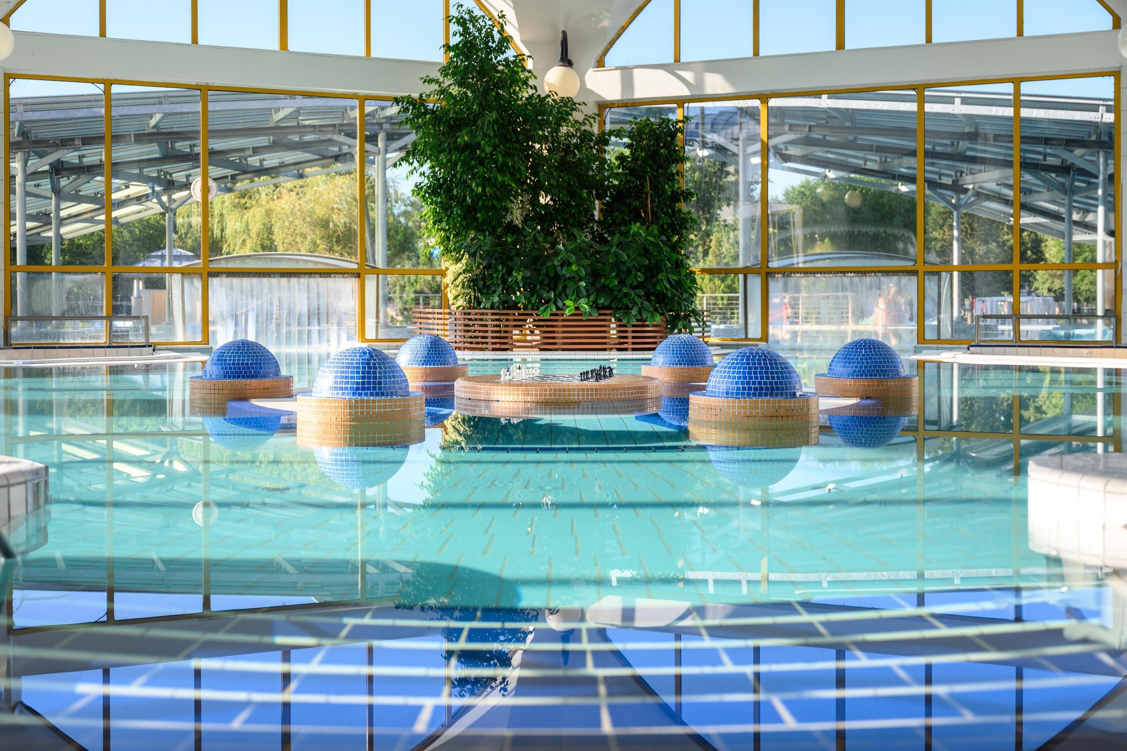 Una piscina interior con paredes de vidrio transparente, decoraciones esféricas azules y una zona central con plantas.