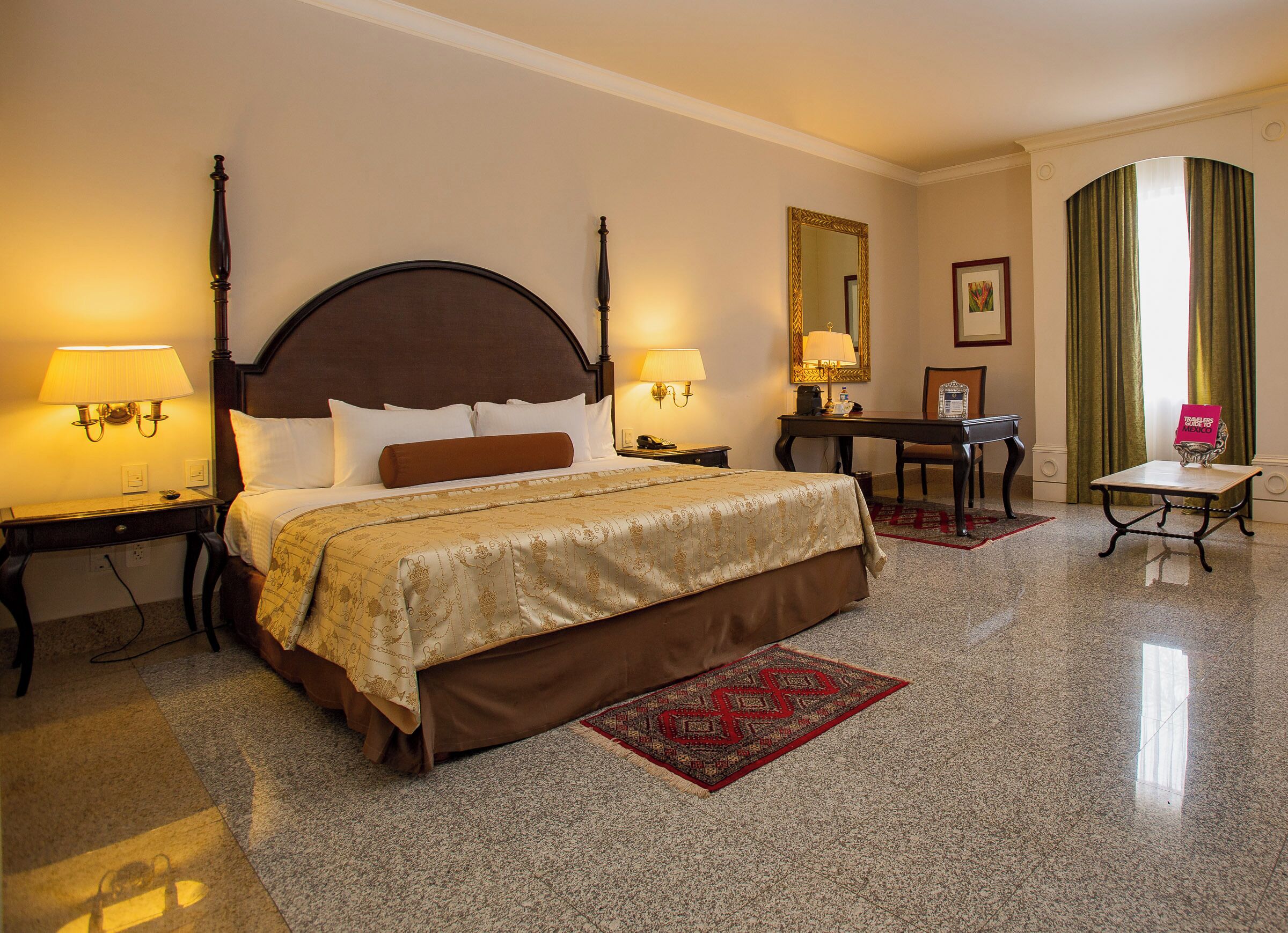 A hotel room with a large bed, two bedside tables with lamps, a desk with a chair, a mirror, and a window with curtains.