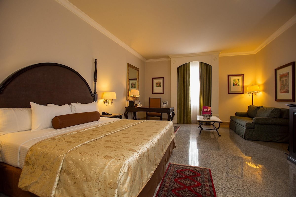 A hotel room with a large bed, a desk, a chair, a sofa, and a small table.