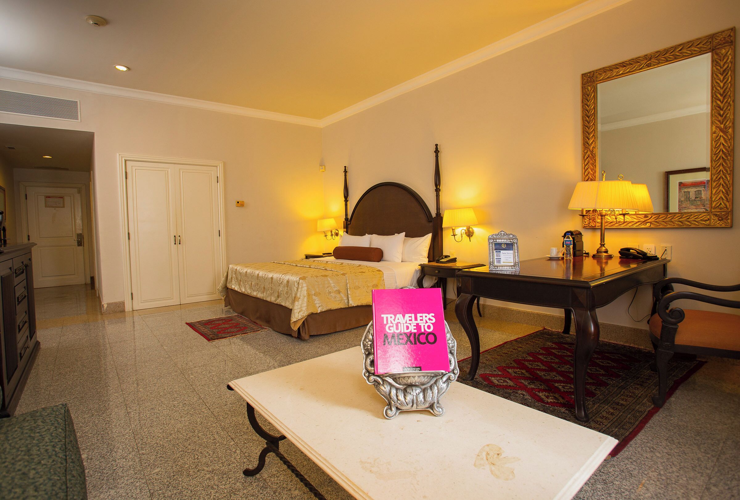 A hotel room with a bed, bedside lamps, a desk with a lamp, a mirror, and a 'Travelers Guide to Mexico' on a stand.