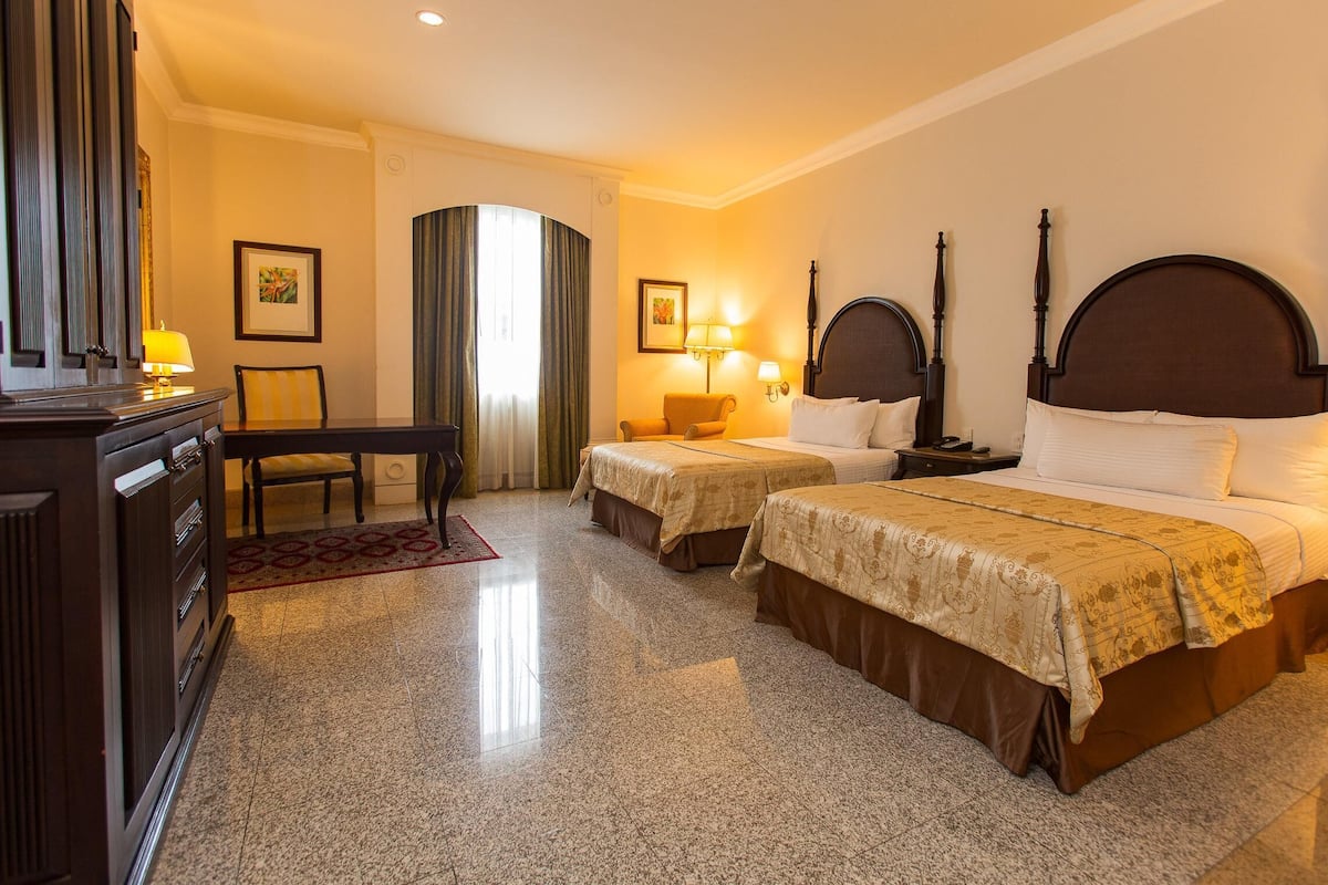 A hotel room with two beds, a desk, a chair, lamps, and framed artwork on the walls.