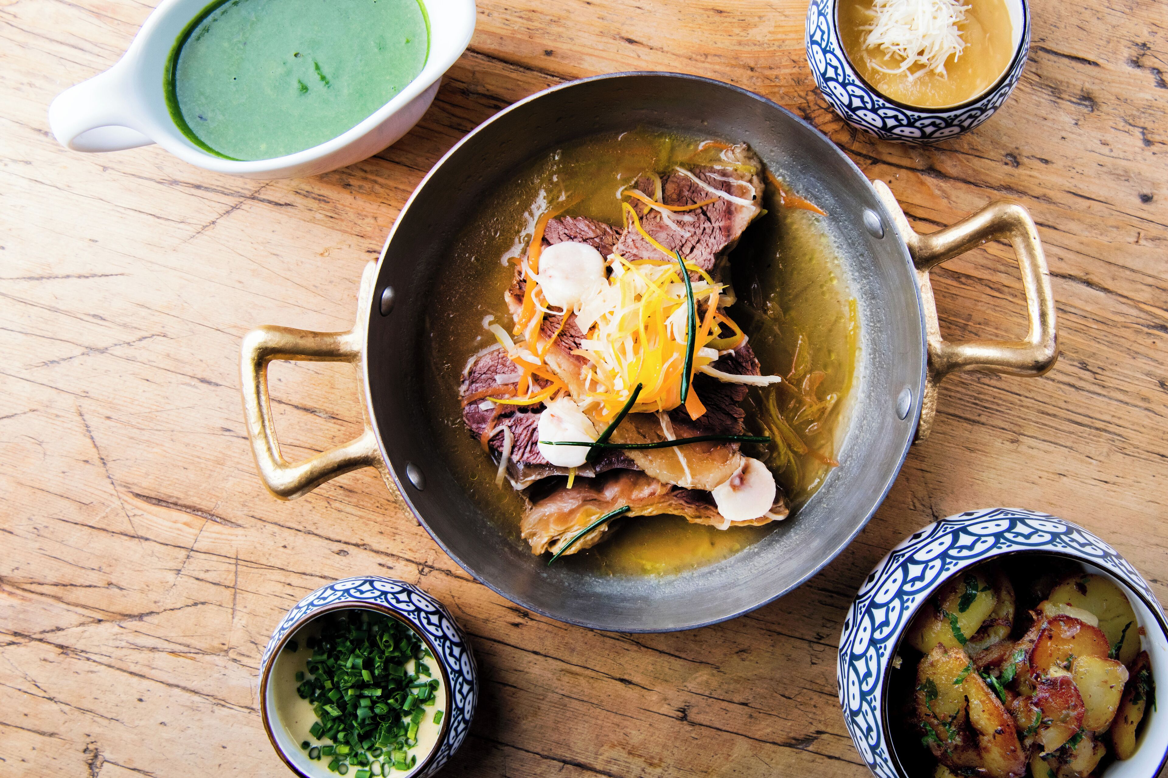 A pan with meat and vegetables, surrounded by bowls of sauces and garnishes on a wooden table.