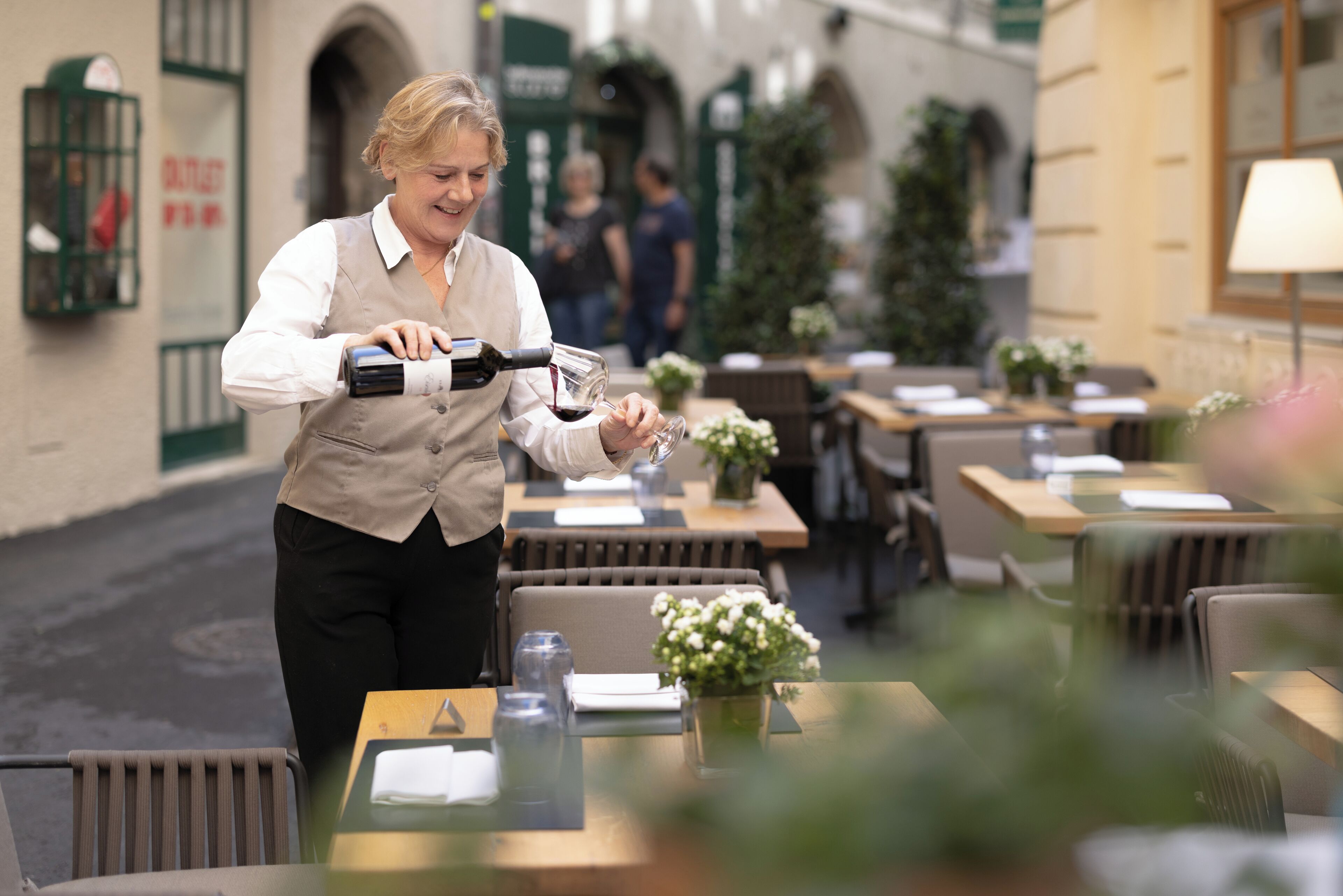 A waiter pouring wine into a glass at an outdoor café with tables and chairs, and a building with green shutters in the background.