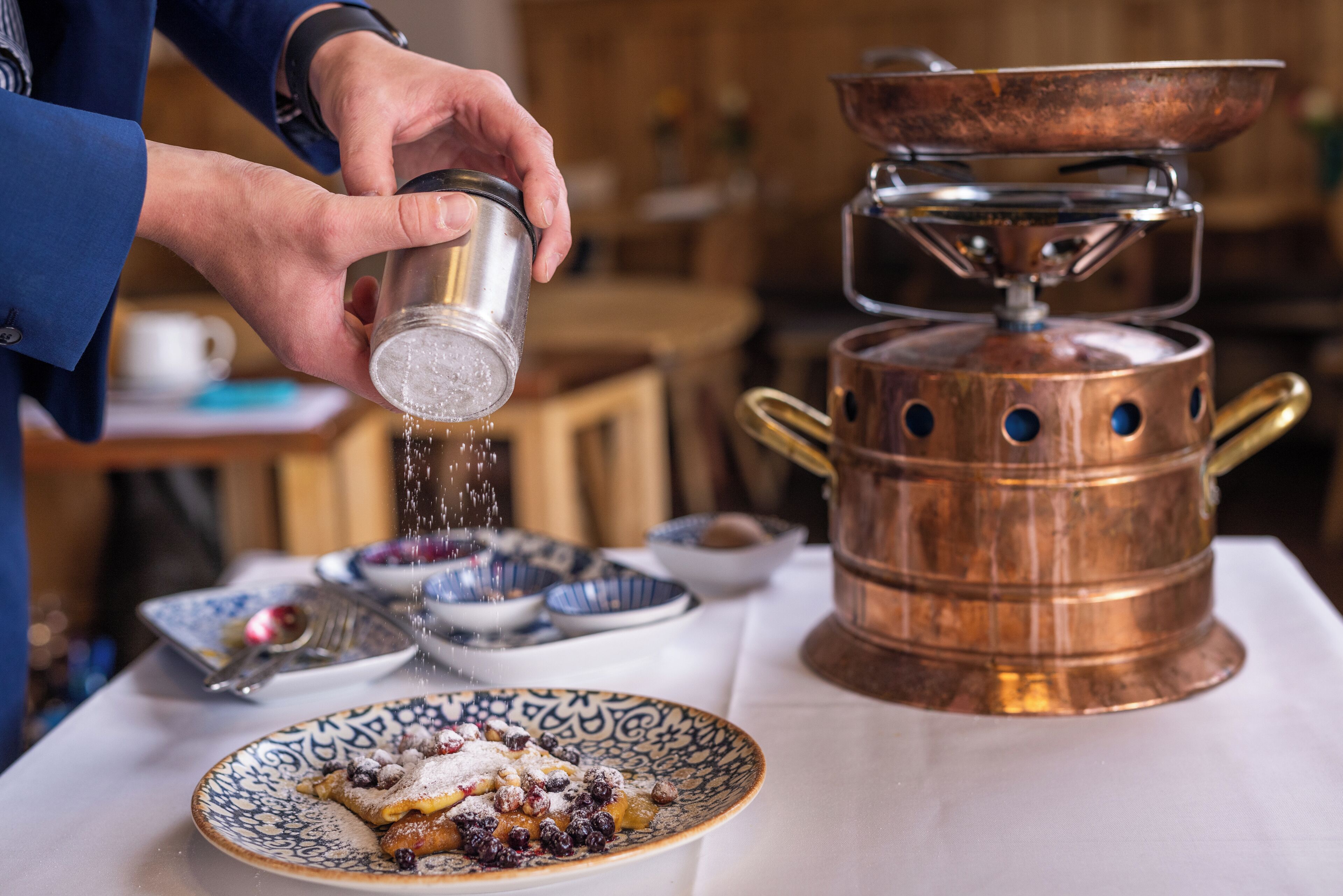 A person sprinkling sugar over a dessert on a plate, with a copper stove and bowls in the background.