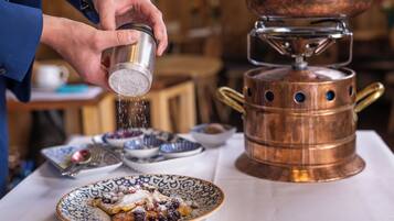 A person sprinkling sugar over a dessert on a plate, with a copper stove and bowls in the background.