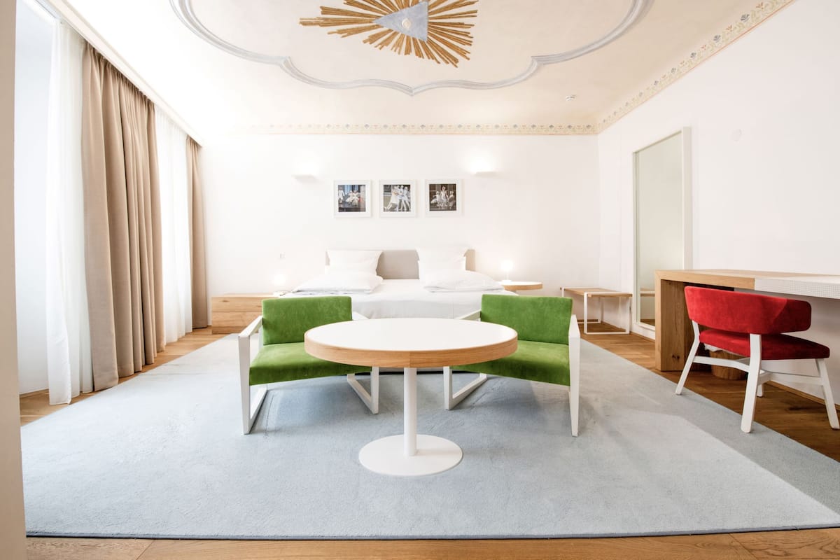 Ein modernes Hotelzimmer mit rundem Tisch, grünen und roten Stühlen, einem Bett mit weißer Bettwäsche und einem großen Spiegel.