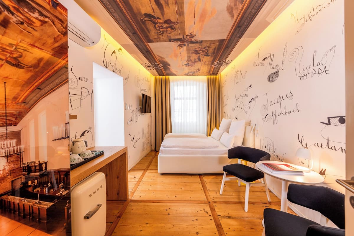 Ein Hotelzimmer mit einem Bett, einem Schreibtisch mit altem Kühlschrank, einem Sessel, einem kleinen Tisch und einer Wand mit Namen und Kritzeleien.