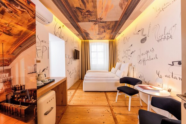 A hotel room with a bed, a desk with a vintage refrigerator, a chair, a small table, and a wall with various names and doodles.