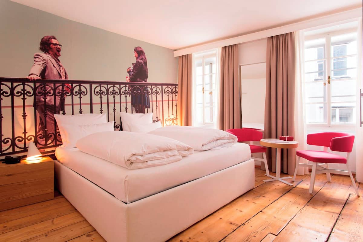 Ein Hotelzimmer mit einem großen Bett, einem kleinen Tisch, zwei roten Stühlen und einem Balkon mit Geländer.