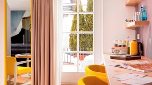 Een moderne kamer met een groot raam, een gele stoel, een tafel met een waterkoker en een boekenkast met potjes.