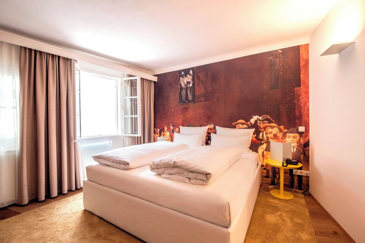 Ein Hotelzimmer mit einem großen Bett, einem Schreibtisch mit Lampe und einem Wandgemälde.
