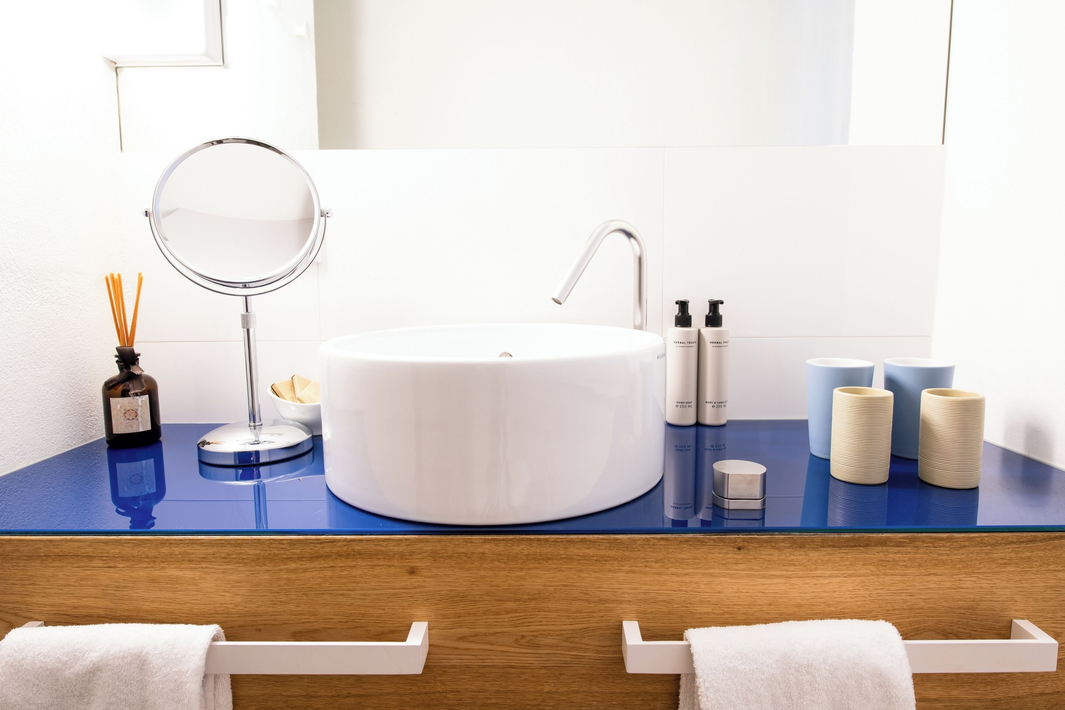 Ein Badezimmer mit einem weißen Waschbecken, einem runden Spiegel und einer blauen Ablagefläche. Auf der Ablagefläche befinden sich zwei Seifenflaschen und zwei Kerzen.