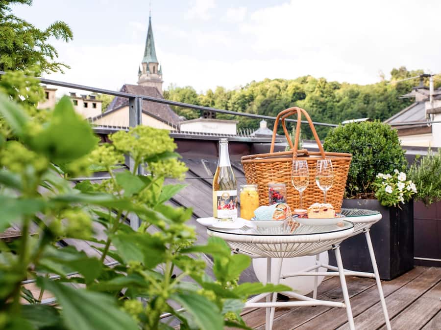 Ein Balkon mit einem für ein Picknick gedeckten Tisch, darunter eine Flasche, ein Korb und Speisen.