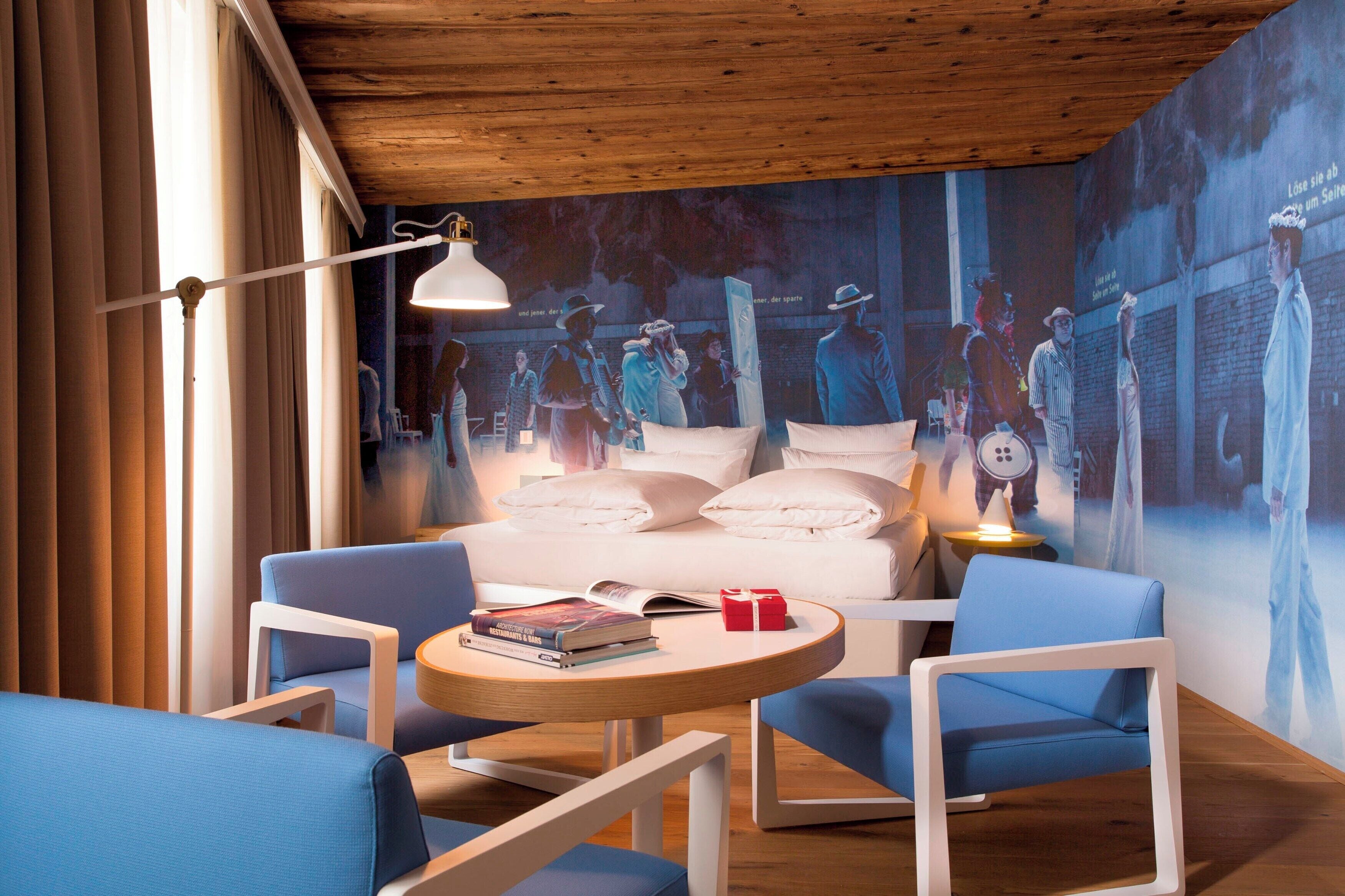 Una habitación de hotel moderna con una cama grande, dos sillones azules, una mesita redonda y un mural en la pared.