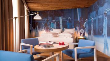 Una habitación de hotel moderna con una cama grande, dos sillones azules, una mesita redonda y un mural en la pared.
