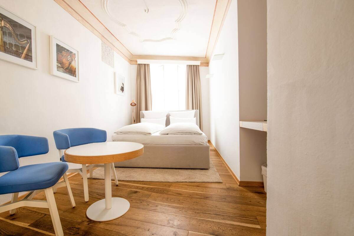 Ein moderner Raum mit einem Bett, zwei blauen Stühlen, einem runden Tisch und Holzboden.