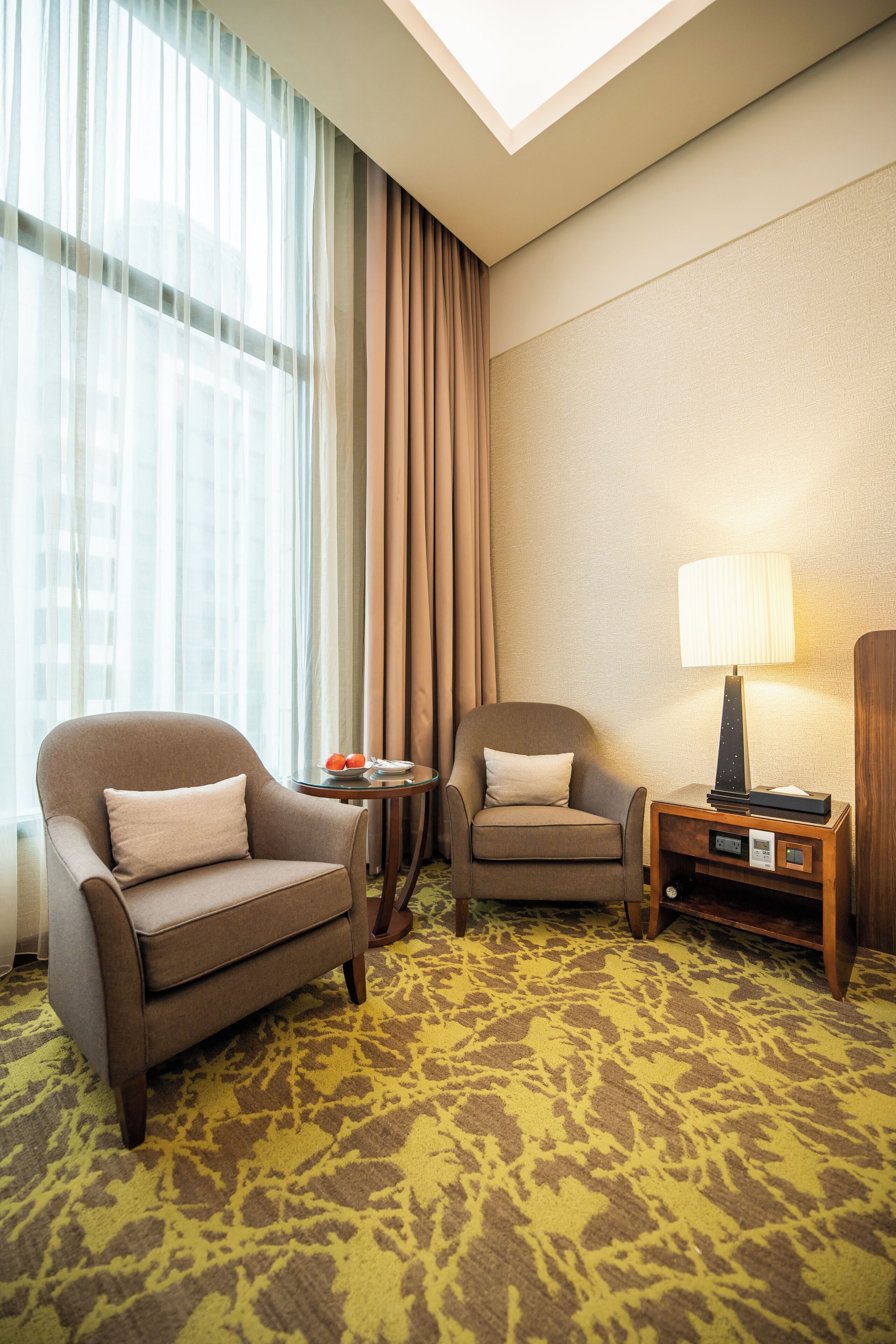 A hotel room with two armchairs, a small table with a lamp, and a view of the city through sheer curtains.