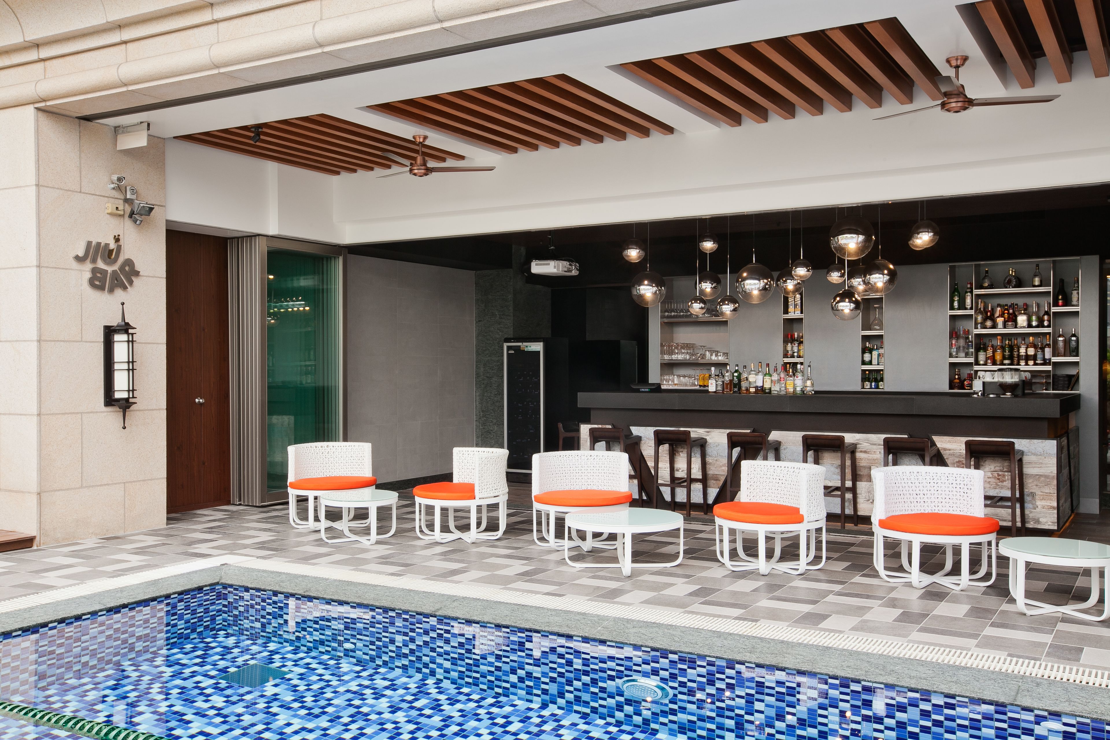 A poolside area with white wicker chairs and orange cushions, a bar with shelves of bottles, and a ceiling fan.