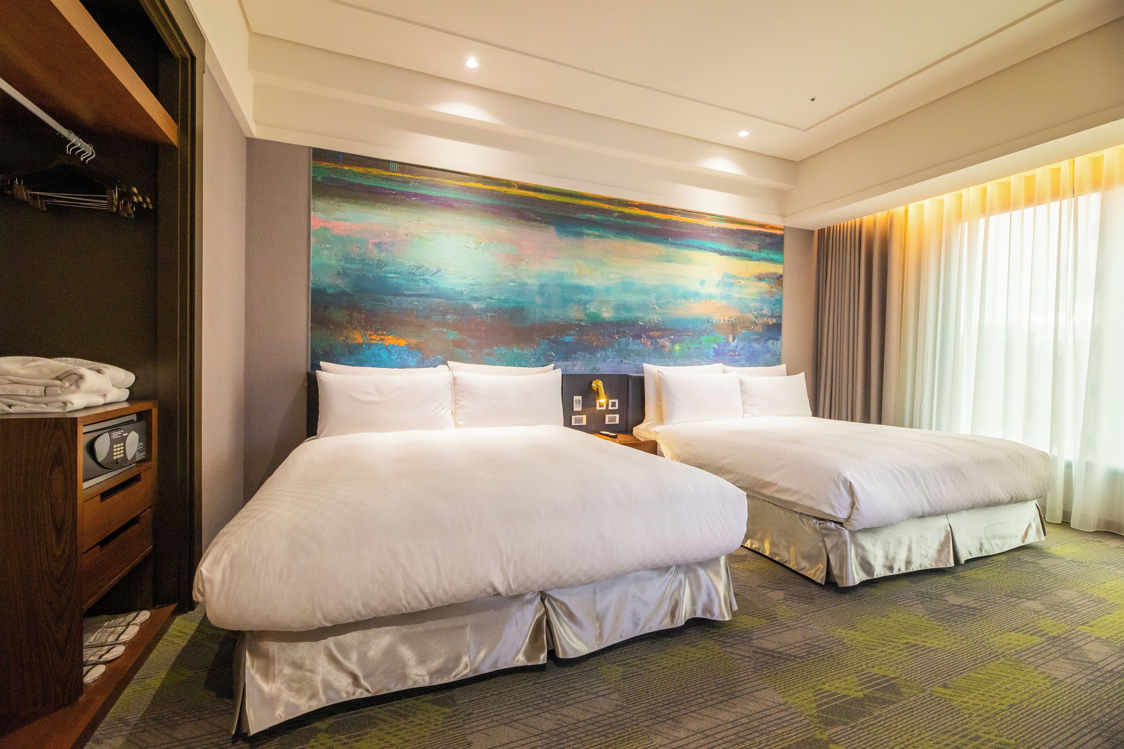 A hotel room with two beds, a large abstract painting on the wall, a wooden dresser, and a window with curtains.