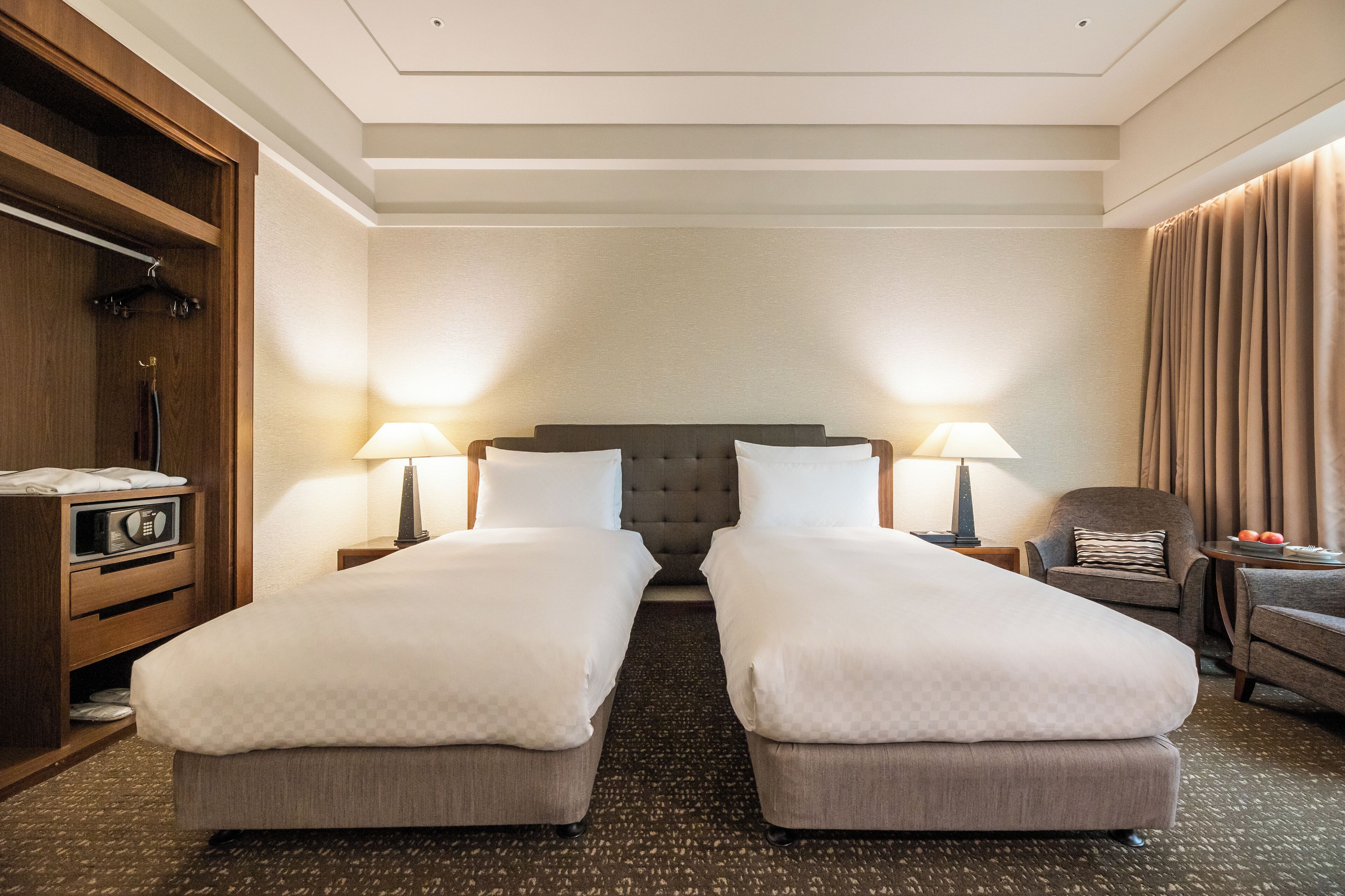 A hotel room with two beds, a wooden wardrobe, a desk with a lamp, and a chair.