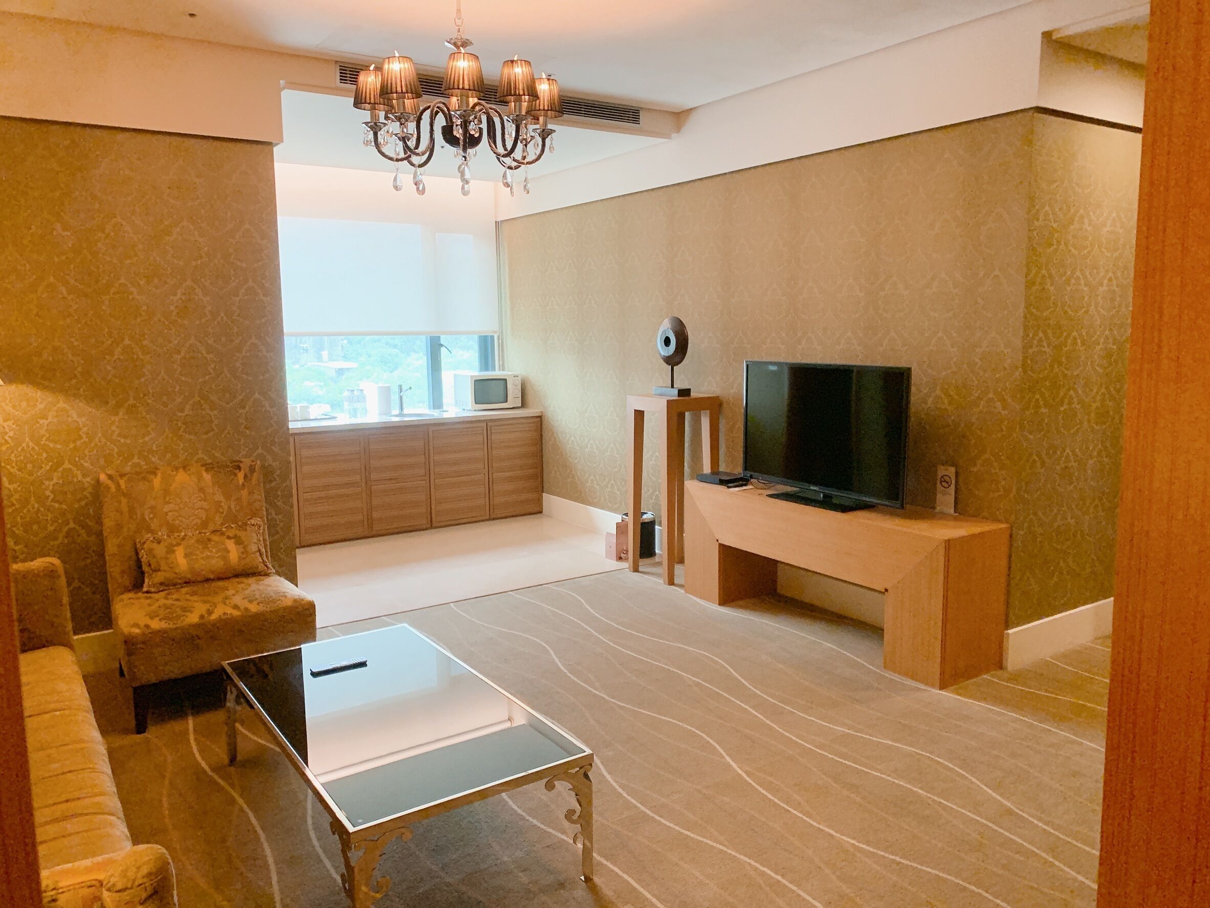 A modern living room with a sofa, a glass coffee table, a TV on a wooden stand, a chandelier, and a view through a window.
