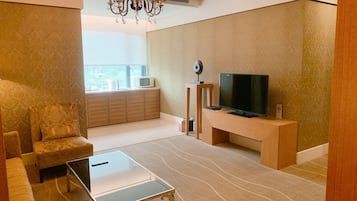 A modern living room with a sofa, a glass coffee table, a TV on a wooden stand, a chandelier, and a view through a window.