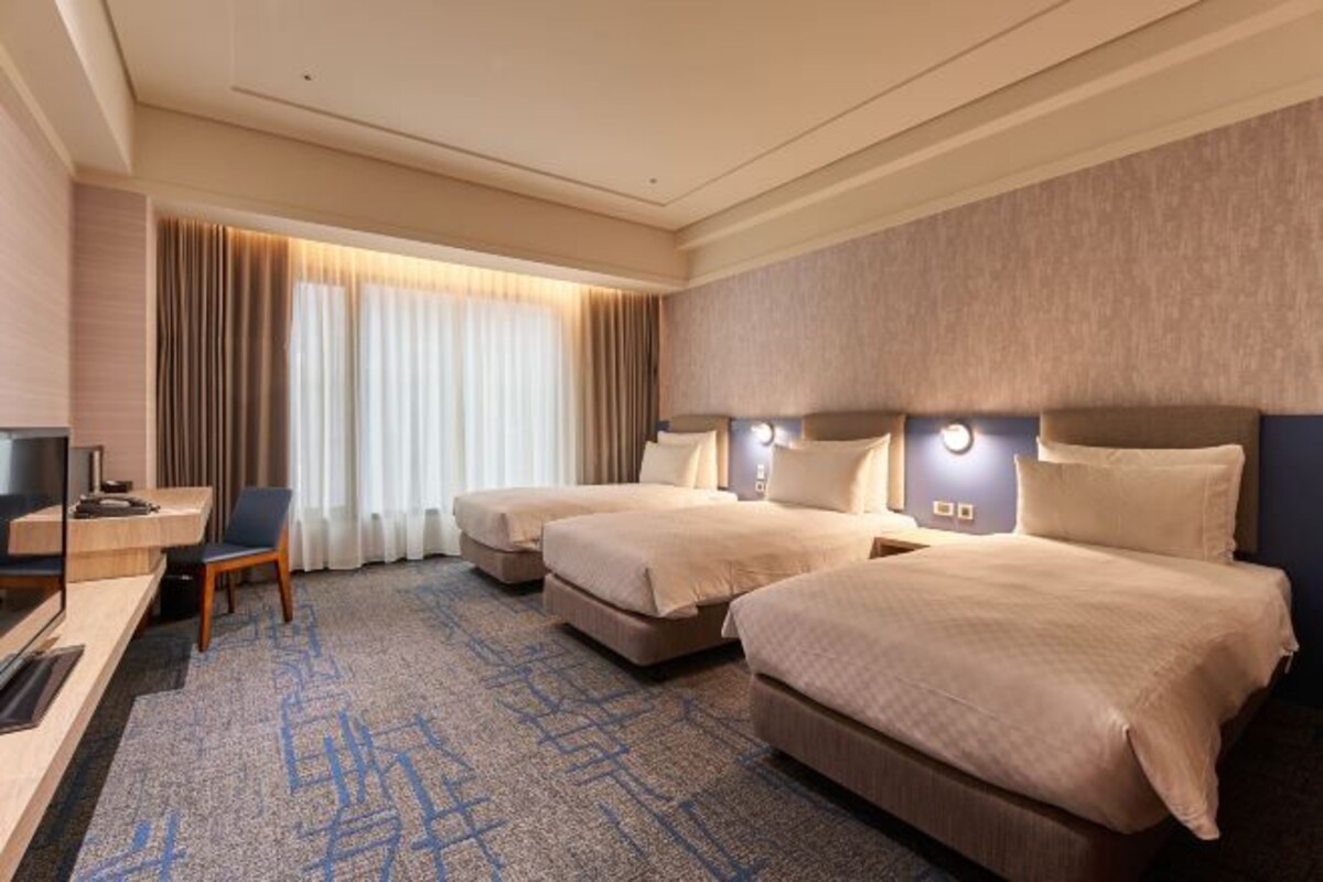 A hotel room with two beds, a desk, a chair, and a large window with curtains.